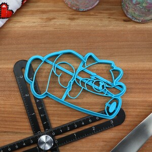 May include: A blue plastic cookie cutter shaped like a panda bear. The cutter is lying on a wooden surface with a ruler and a spatula.