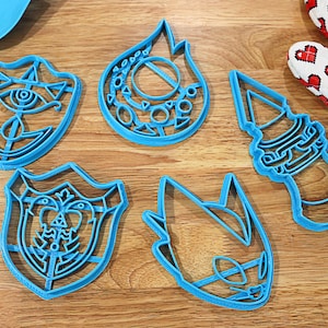 May include: Five blue plastic cookie cutters shaped like a flame, a shield, a face, a tower, and a head. The cutters are on a wooden surface.