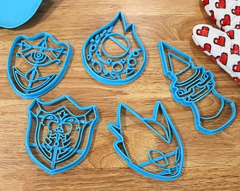 Puede incluir: Cinco cortadores de galletas de plástico azul con forma de llama, escudo, cara, torre y cabeza. Los cortadores están sobre una superficie de madera.