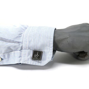 May include: A pair of cufflinks with a silver finish and a geometric design. The cufflinks are on a blue and white striped shirt cuff.