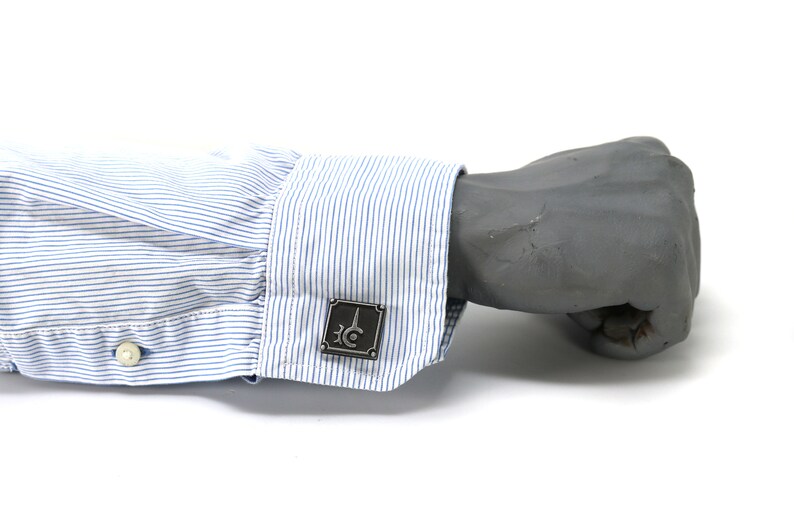 May include: A pair of cufflinks with a silver finish and a geometric design. The cufflinks are on a blue and white striped shirt cuff.