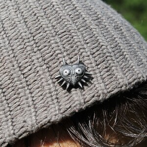 May include: A grey knitted beanie with a silver pin featuring a stylised heart with a face and spikes.