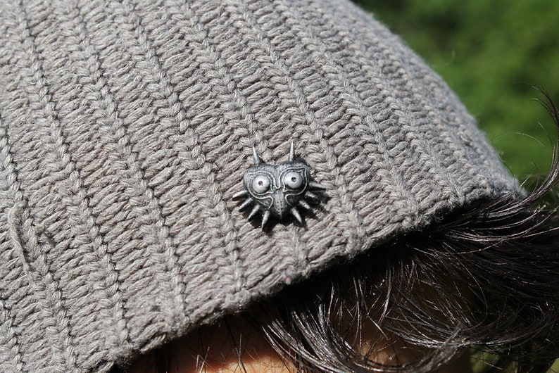 May include: A grey knitted beanie with a silver pin featuring a stylised heart with a face and spikes.