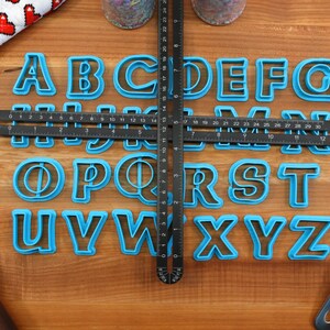 May include: A set of blue plastic alphabet cookie cutters on a wooden surface. The cutters are arranged in alphabetical order. A black ruler is placed on top of the cutters.