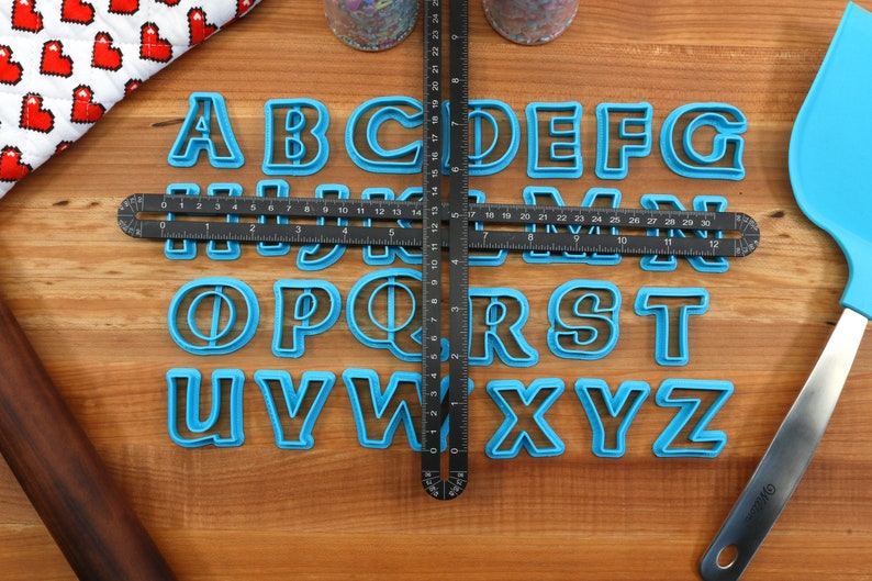 May include: A set of blue plastic alphabet cookie cutters on a wooden surface. The cutters are arranged in alphabetical order. A black ruler is placed on top of the cutters.