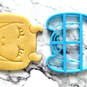 May include: A blue plastic cookie cutter shaped like a sleeping unicorn, next to a yellow sugar cookie cut with the same shape.