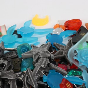 May include: A collection of colorful plastic and resin charms in various shapes, including stars, suns, moons, and abstract designs. The charms are in shades of blue, green, red, orange, and purple.