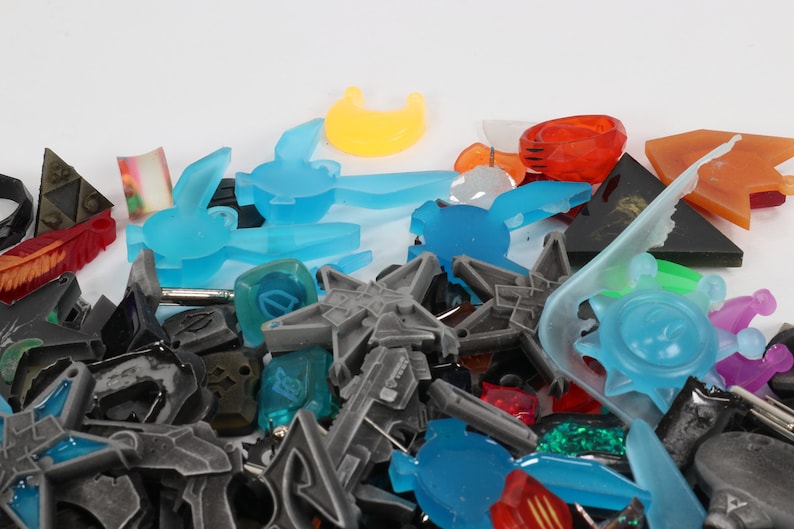 May include: A collection of colorful plastic and resin charms in various shapes, including stars, suns, moons, and abstract designs. The charms are in shades of blue, green, red, orange, and purple.