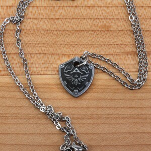 May include: A silver chain necklace with a silver pendant shaped like a shield with a black and white design of a bird inside a triangle.