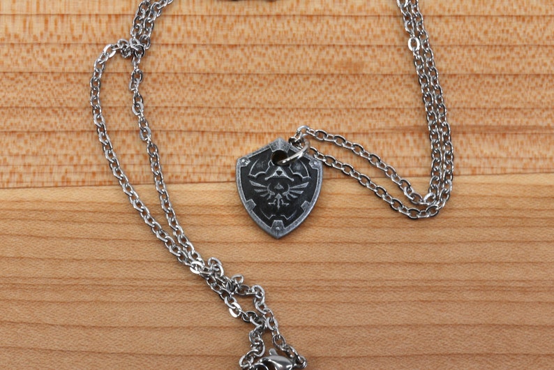 May include: A silver chain necklace with a silver pendant shaped like a shield with a black and white design of a bird inside a triangle.