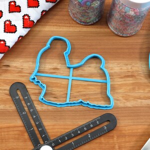 May include: Blue plastic cookie cutter shaped like a sitting dog. The cutter is on a wooden surface with a ruler and a spatula.