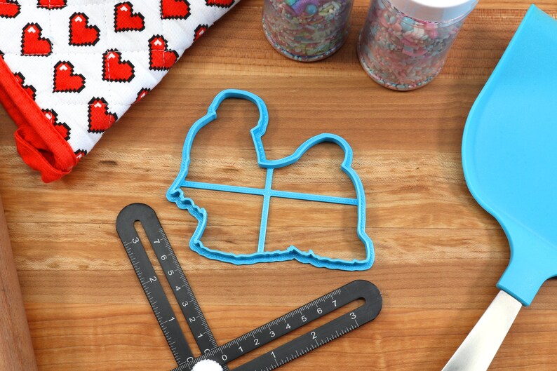 May include: Blue plastic cookie cutter shaped like a sitting dog. The cutter is on a wooden surface with a ruler and a spatula.