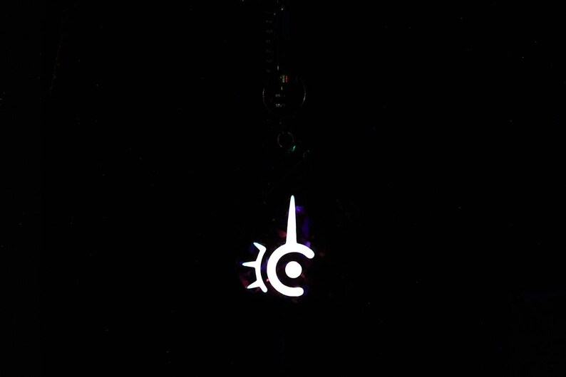 May include: A white pendant with a unique, stylized design, hanging against a black background. The pendant features a central circle with a vertical spike and a gear-like shape on the left side. The design glows brightly.