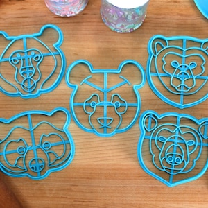 May include: Five blue plastic cookie cutters shaped like bear faces. The cutters have a grid pattern inside the bear shapes. The cutters are on a wooden surface.