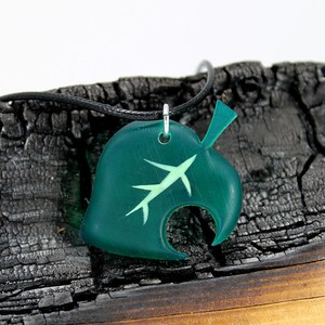 May include: A green leaf-shaped pendant necklace with a white outline of a leaf inside. The necklace is on a black cord and is resting on a piece of burnt wood.