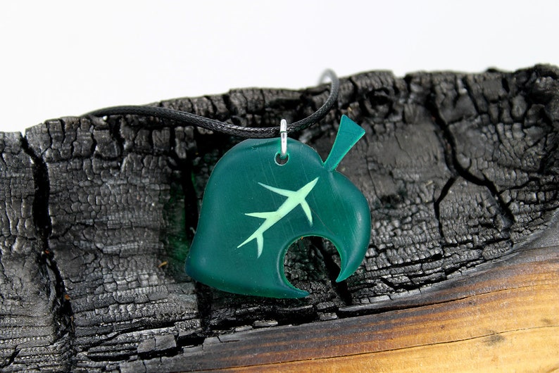 May include: A green leaf-shaped pendant necklace with a white outline of a leaf inside. The necklace is on a black cord and is resting on a piece of burnt wood.
