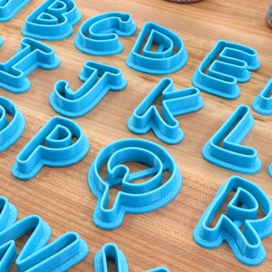 Op de afbeelding: Een set blauwe plastic alfabetkoekjesnijders. De snijders zijn gerangschikt op een houten oppervlak.