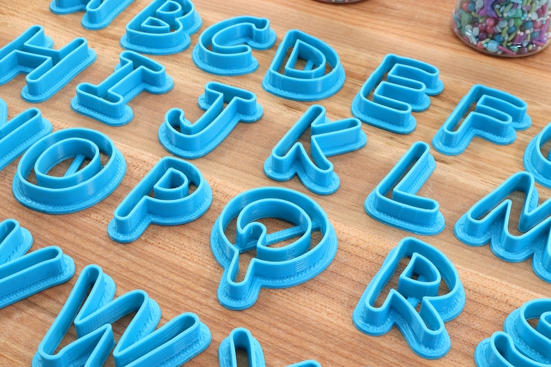 Op de afbeelding: Een set blauwe plastic alfabetkoekjesnijders. De snijders zijn gerangschikt op een houten oppervlak.