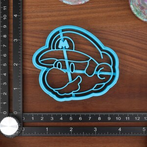 May include: A blue plastic cookie cutter shaped like a cartoon character with a mustache and a hat. The character is wearing a hat with an 'M' on it.
