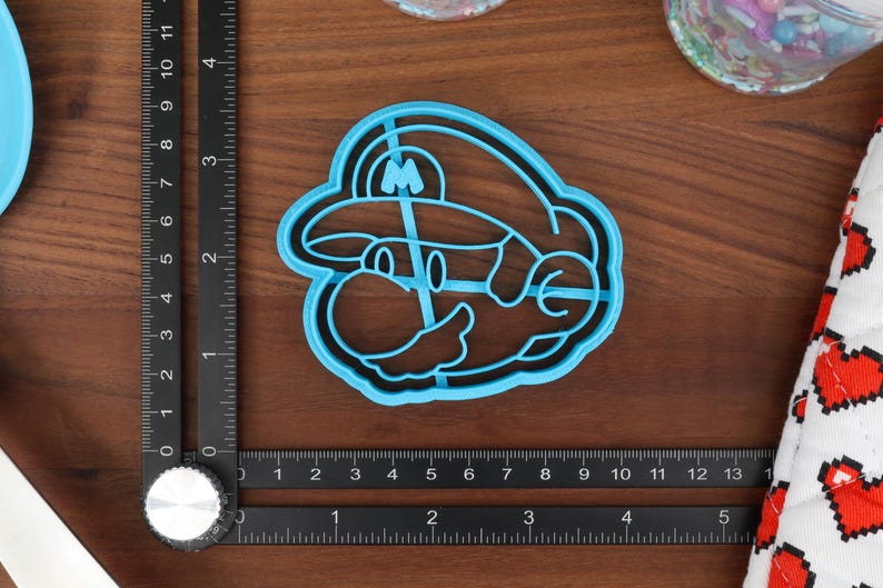 May include: A blue plastic cookie cutter shaped like a cartoon character with a mustache and a hat. The character is wearing a hat with an 'M' on it.