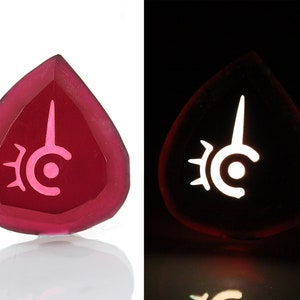 May include: A pink glass pendant with a white symbol of a crescent moon with a spike on top, surrounded by a sunburst design. The pendant glows in the dark.