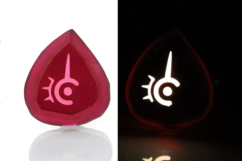 May include: A pink glass pendant with a white symbol of a crescent moon with a spike on top, surrounded by a sunburst design. The pendant glows in the dark.