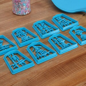 May include: A collection of eight turquoise cookie cutters, each shaped as a rectangle with Chinese characters. The cutters are arranged on a wooden surface, with jars of colorful sprinkles and a blue spatula in the background. The cutters are for baking.