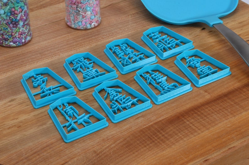 May include: A collection of eight turquoise cookie cutters, each shaped as a rectangle with Chinese characters. The cutters are arranged on a wooden surface, with jars of colorful sprinkles and a blue spatula in the background. The cutters are for baking.