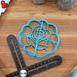 May include: A blue plastic cookie cutter shaped like a rose. The cutter is on a wooden surface with a ruler and other baking tools.