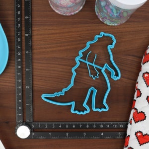 May include: Blue plastic cookie cutter shaped like a dinosaur, measuring approximately 5 inches tall, on a wooden surface with a ruler and a heart-patterned fabric.