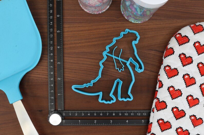 May include: Blue plastic cookie cutter shaped like a dinosaur, measuring approximately 5 inches tall, on a wooden surface with a ruler and a heart-patterned fabric.