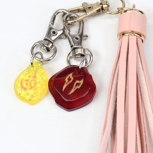 Puede incluir: Un llavero de borla de cuero rosa con dos dijes. Un dije es amarillo con un dise&ntilde;o dorado y el otro es rojo con un dise&ntilde;o dorado.