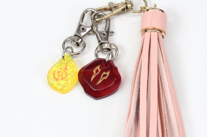 Puede incluir: Un llavero de borla de cuero rosa con dos dijes. Un dije es amarillo con un dise&ntilde;o dorado y el otro es rojo con un dise&ntilde;o dorado.