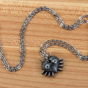 May include: A silver chain necklace with a Majora's Mask pendant. The pendant is a heart shape with a detailed design of the mask, including eyes, a nose, and teeth. The mask is made of metal and has a dark gray finish.