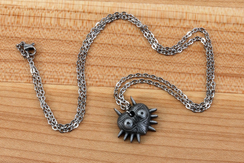 May include: A silver chain necklace with a Majora's Mask pendant. The pendant is a heart shape with a detailed design of the mask, including eyes, a nose, and teeth. The mask is made of metal and has a dark gray finish.