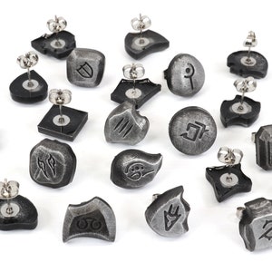 Puede incluir: Colecci&oacute;n de pendientes de bot&oacute;n plateados con varios s&iacute;mbolos y formas grabadas. Los pendientes tienen una base gris oscuro y postes de acero inoxidable. Los dise&ntilde;os incluyen formas geom&eacute;tricas, runas y otros patrones abstractos.