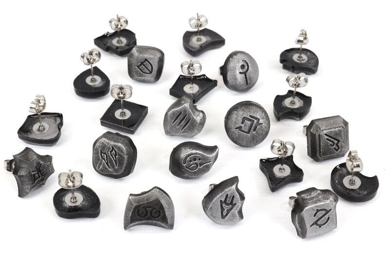 Puede incluir: Colecci&oacute;n de pendientes de bot&oacute;n plateados con varios s&iacute;mbolos y formas grabadas. Los pendientes tienen una base gris oscuro y postes de acero inoxidable. Los dise&ntilde;os incluyen formas geom&eacute;tricas, runas y otros patrones abstractos.