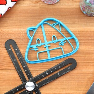 May include: A blue plastic cookie cutter shaped like a cartoon cat with a wide smile and big eyes. The cutter is on a wooden surface with a ruler and a rolling pin.