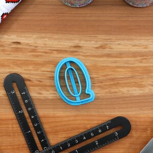 May include: A blue plastic cookie cutter shaped like the letter Q. The cutter is on a wooden surface.