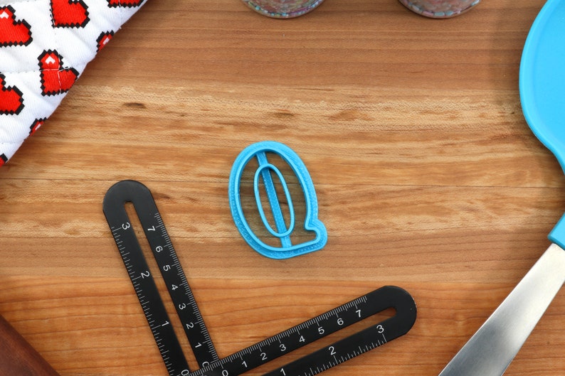 May include: A blue plastic cookie cutter shaped like the letter Q. The cutter is on a wooden surface.