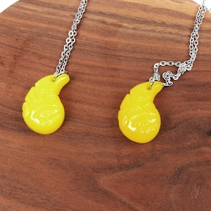 May include: Two yellow teardrop-shaped pendants on silver chains. The pendants have intricate designs and are hanging against a wooden background.