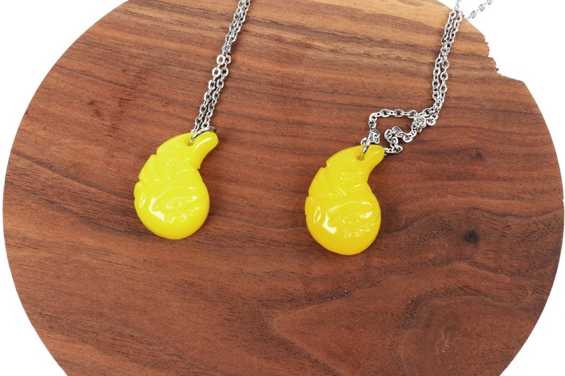 May include: Two yellow teardrop-shaped pendants on silver chains. The pendants have intricate designs and are hanging against a wooden background.