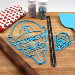 May include: Two blue plastic cookie cutters shaped like a cartoon face with a hat, a rolling pin, a spatula, a ruler, and two jars of sprinkles on a wooden cutting board.
