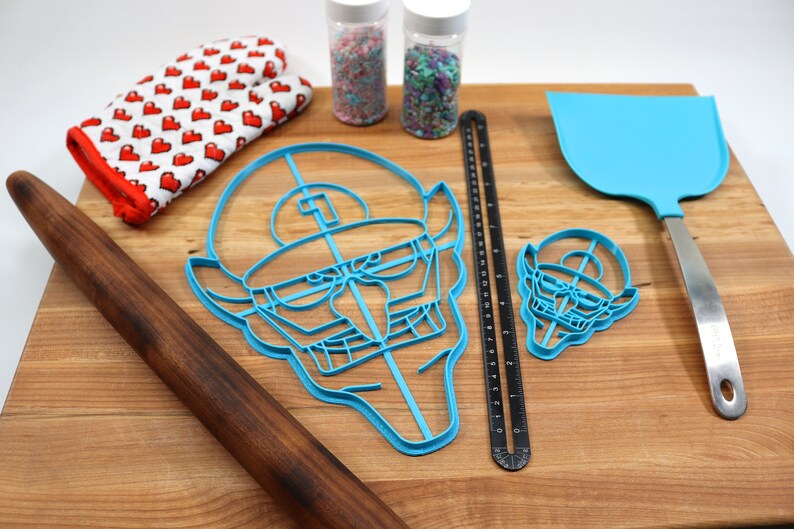 May include: Two blue plastic cookie cutters shaped like a cartoon face with a hat, a rolling pin, a spatula, a ruler, and two jars of sprinkles on a wooden cutting board.