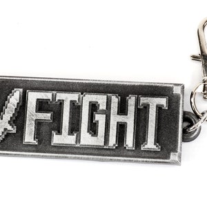 May include: A silver metal keychain with a black rectangular tag that says "FIGHT" in white pixelated letters. A silver sword icon is to the left of the text. The keychain has a silver swivel clasp.