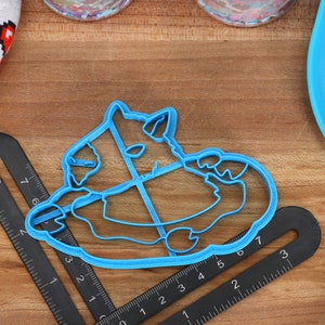 May include: Blue plastic cookie cutter shaped like a cartoon monster with a big belly and a wide grin. The cutter is on a wooden surface with a ruler.
