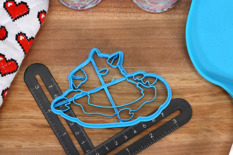 May include: Blue plastic cookie cutter shaped like a cartoon monster with a big belly and a wide grin. The cutter is on a wooden surface with a ruler.