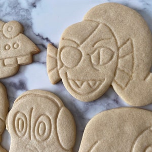 K&ouml;nnte beinhalten: F&uuml;nf einfache Zuckerkekse, die mit verschiedenen Comicfiguren verziert sind. Die Kekse liegen auf einer wei&szlig;en Marmorfl&auml;che.