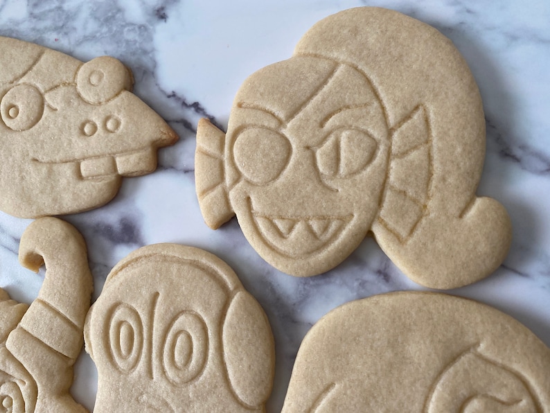 K&ouml;nnte beinhalten: F&uuml;nf einfache Zuckerkekse, die mit verschiedenen Comicfiguren verziert sind. Die Kekse liegen auf einer wei&szlig;en Marmorfl&auml;che.