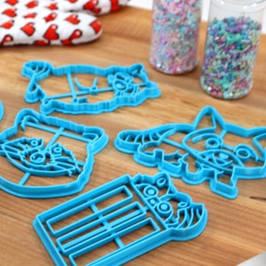 Puede incluir: Un juego de seis cortadores de galletas de pl&aacute;stico azul con forma de mapaches. Los cortadores est&aacute;n sobre una superficie de madera con chispas en frascos en el fondo.